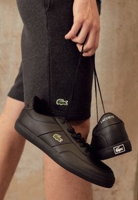 Une main tenant des baskets Lacoste noires par les lacets, une personne portant un short gris foncé avec le logo crocodile Lacoste sur le côté.