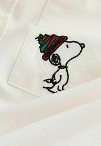 Gestickter Cartoon-Hund mit einer rot-grün gestreiften Wintermütze auf weißem Stoff mit einem gestickten Taschen-Detail.