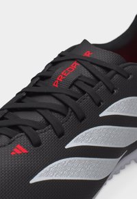 Μαύρο παπούτσι ποδοσφαίρου με μαύρα κορδόνια, κόκκινο κείμενο "PREDATOR" στη γλώσσα και ασημένιες υφασμένες ρίγες στο πλάι.