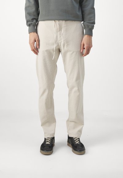 Beige textured pants with an elastic waistband and drawstring. Paired with black sneakers featuring a rubber sole and cream laces.