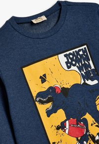 Blauwe sweatshirt met een geribde halslijn, voorzien van een dinosaurussenafbeelding die een voetbal vasthoudt, tegen een oranje achtergrond met zwarte tekst.