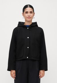 Vrouw in een zwarte hooded jack met zilveren drukknoopjes en voorzakken, gecombineerd met donkerkleurige losse broek, tegen een effen achtergrond.