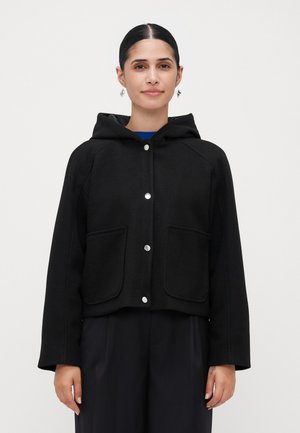 Woman wearing a black hooded jacket with silver snap buttons and front pockets, paired with dark loose-fitting pants against a plain background.