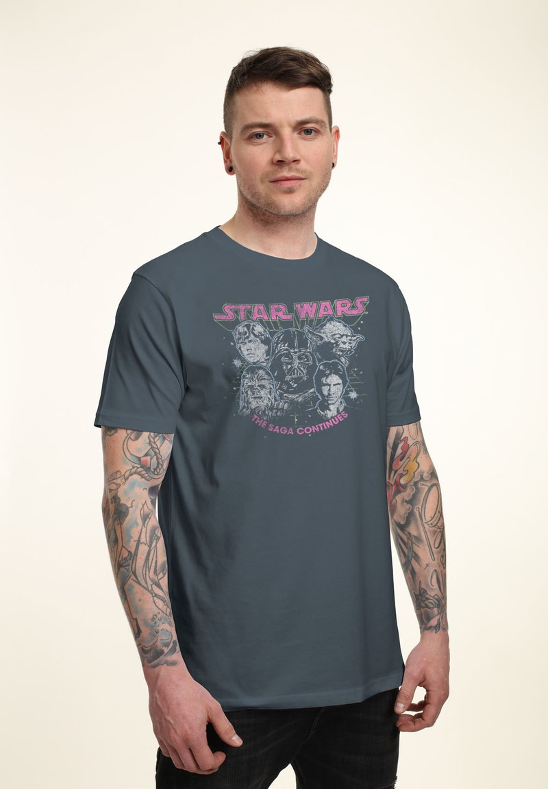 Homme tatoué portant un t-shirt Star Wars sombre avec des personnages et le texte "La saga continue", debout devant un fond uni.