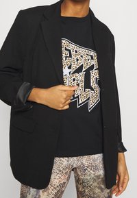 Blazer negro con tejido texturizado, combinado con una camiseta negra con letras de estampado de leopardo y leggings con patrón de serpiente.