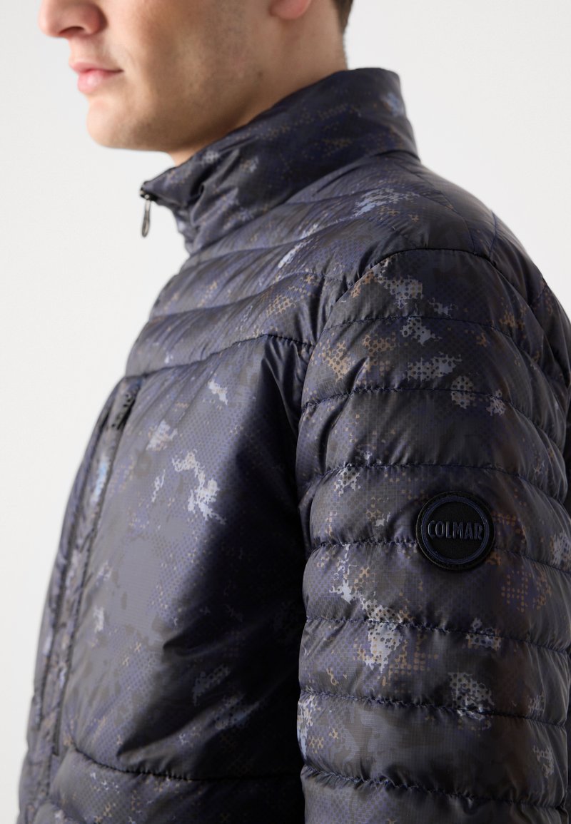 Granatowa kurtka puchowa z geometrycznym wzorem, z wysokim kołnierzem, zapięciem na zamek oraz okrągłym logo Colmar na lewym rękawie.