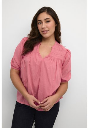 Rosa Bluse mit kurzen Puffärmeln, V-Ausschnitt und bestickten Details an der Passe; kombiniert mit dunklen Jeans. Glatte Stoffstruktur.