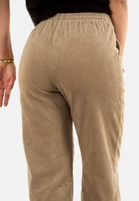 Pantalons en velours côtelé beige avec des côtes verticales, une taille élastique et des poches latérales. Le tissu a une finition douce et texturée.