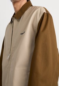 Beige en bruin gekleurd blokjasje met rits, puntige kraag en subtiel logo op de borst. Gemaakt van duurzaam materiaal.