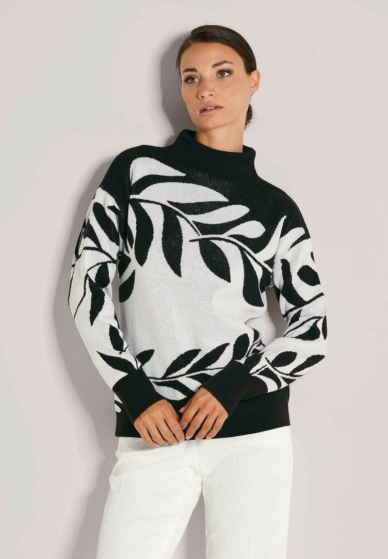 Frau trägt einen schwarz-weißen Pullover mit großen Blattmustern und weiße Hosen, steht vor einem schlichten hellen Hintergrund.