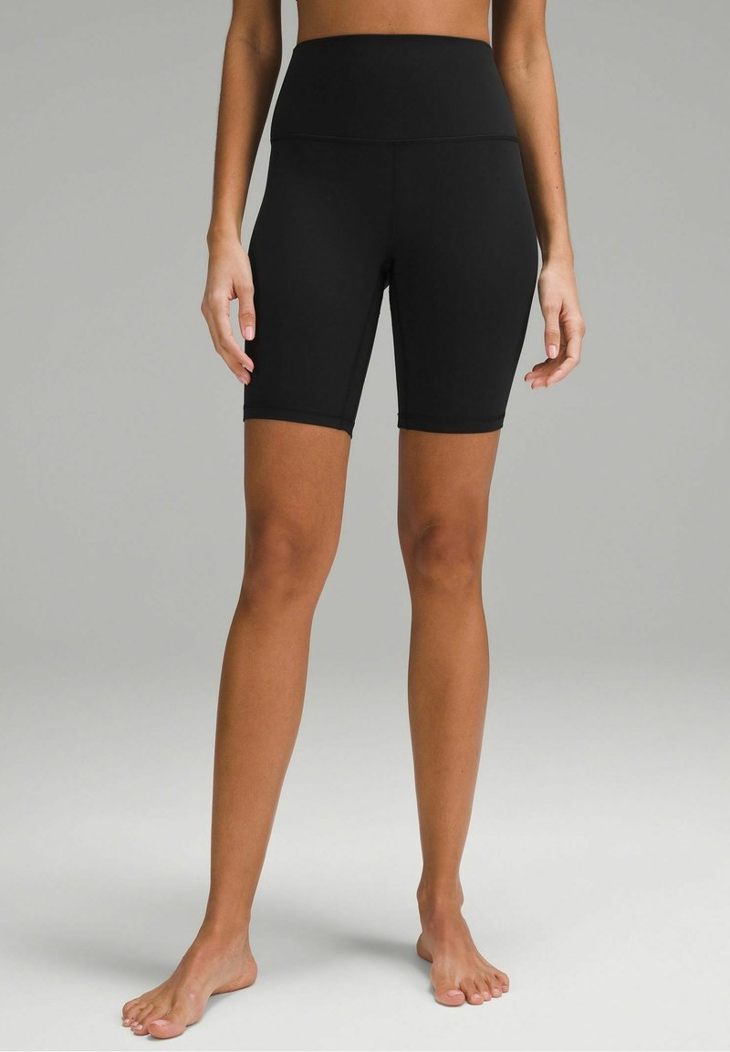 Femme portant un short noir ajusté, taille haute, arrivant au genou, debout pieds nus devant un fond gris uni.