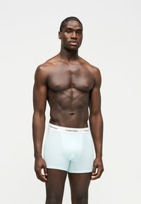 Hombre musculoso de pie, vistiendo bóxers Calvin Klein azul claro contra un fondo blanco liso, mirando hacia adelante con expresión neutral.