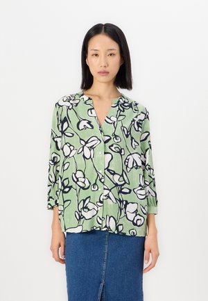 Chemise à boutonner en tissu vert clair avec un motif floral noir et blanc. Présente un col sans col et des manches trois-quarts.