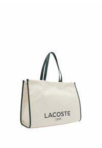Sac fourre-tout en toile de couleur crème avec des bordures et des poignées vert foncé. L'avant présente "LACOSTE 1933" en lettres majuscules imprimées. Forme rectangulaire.