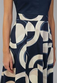 Navy-Kleid mit tailliertem Oberteil und plissiertem Rock, versehen mit einem cremefarbenen und marineblauen kreisförmigen Muster, aus geschmeidigem Stoff mit leichtem Glanz.