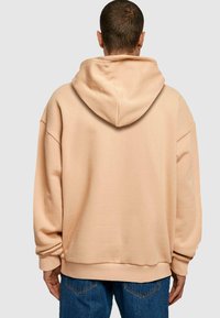 Beige oversized hoodie med snørret hætte, ribbede mansjetter og kant. Glat tekstur og afslappet design. Bæres over blå jeans.