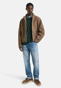 Braune Fischgrät-Zip-Up-Jacke über einem grünen Polo, kombiniert mit hellblauen Jeans und braunen Sneakers mit cremefarbenen Sohlen. Einfaches und lässiges Design.