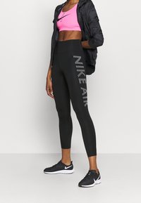 Žena v černých legínách Nike Air, růžovém sportovním podprsenku, černých sportovních botách a černé bundě má ruce v kapsách.