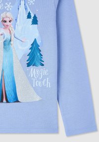 Elsa z Krainy Lodu rzucająca zaklęcie śnieżne z płatkami śniegu, sosnowymi drzewami i śnieżnym zamkiem na jasnoniebieskiej koszulce z długim rękawem.