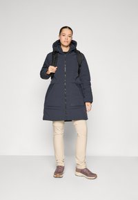 Námornícky puffa kabát so zipsom a kapucňou, obsahujúci dve predné vrecká. Nosený cez béžové nohavice a kombinovaný s hnedými športovými topánkami.