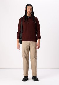 Pull en maille bordeaux, pantalon cargo beige avec poches latérales, et bottes noires à semelle épaisse. Le mannequin porte un sac bandoulière, se tenant devant un fond uni.