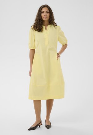 Žena s kučeravými vlasmi nosí pastelovo žlté šaty po kolená s nadýchanými rukávmi a čierne nízke sandále na pätu, stojaca pred jednoduchým pozadím.