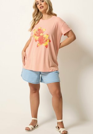Femme blonde posant en t-shirt rose clair avec imprimé floral orange-jaune, short en jean bleu clair et sandales blanches sur fond uni.