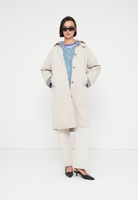 Μπεζ καμπαρντίνα με κουκούλα, διακοσμημένη με ξύλινα κουμπιά, εσωτερική επένδυση από μπλε καπιτονέ ύφασμα και μανσέτες με καρώ σχέδιο, συνδυασμένη με εκρού παντελόνια και μαύρες γόβες.