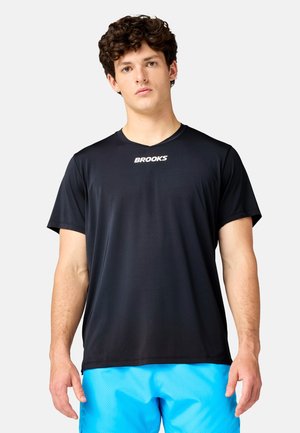 Ung mand med krøllet hår iført en sort Brooks t-shirt og lyseblå sportsshorts mod en ensfarvet baggrund.