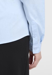 Svijetloplava košulja dugih rukava glatke teksture, s dugmadima na manžetama i zakrivljenim donjim obrubom, u kombinaciji s crnim materijalom.