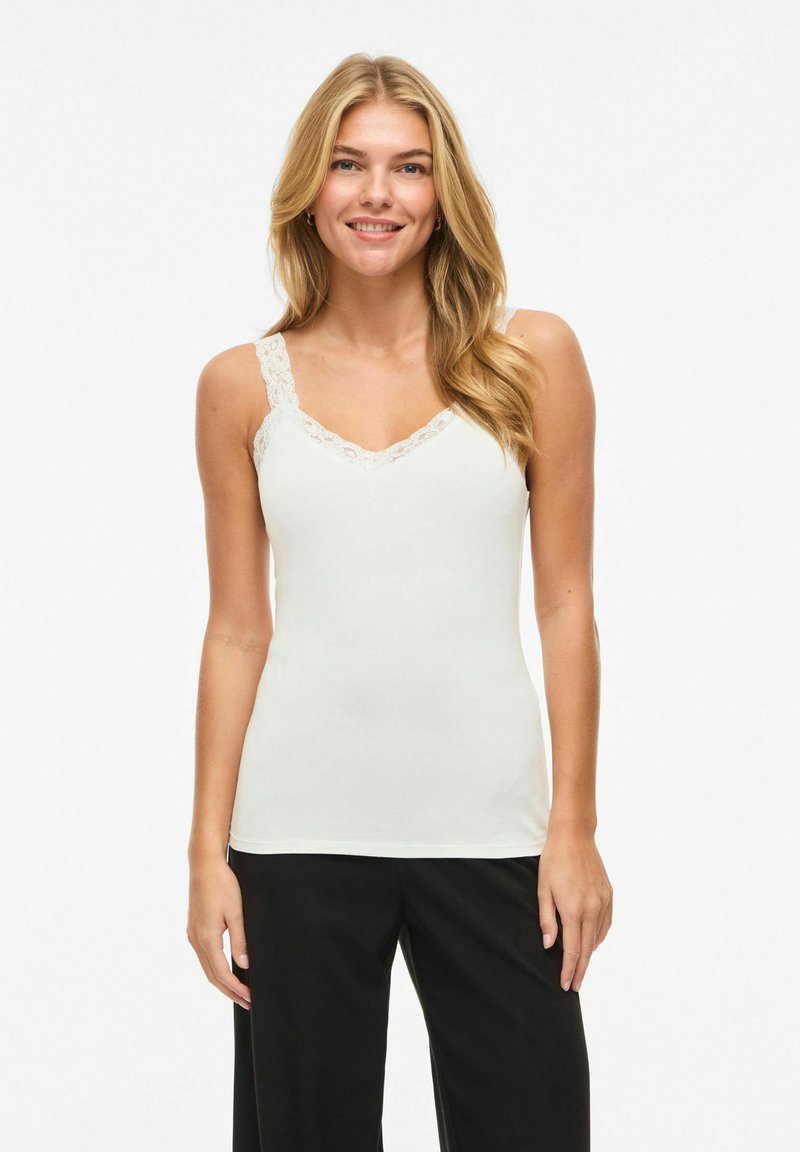 Femme souriante aux longs cheveux blonds portant un haut blanc sans manches orné de dentelle et un pantalon noir, debout devant un fond blanc.