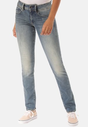 Personne portant un jean slim délavé bleu et des baskets en toile roses claires, se tenant avec une jambe légèrement en avant sur un fond blanc.