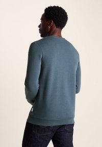Fitted sweatshirt in dark teal, made of soft fabric. Features long sleeves, a crew neckline, and ribbed cuffs and hem. Subtle logo tag at the side.