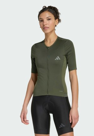 Kvinde iført olivengrøn cykeltrøje med korte ærmer og sorte polstrede cykelshorts stående mod en ensfarvet baggrund.