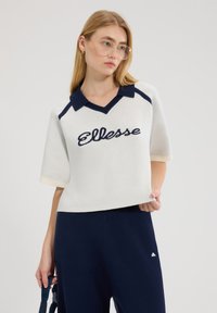 Młoda kobieta ubrana w biały sweter z krótkimi rękawami z niebieskim logo "Ellesse" oraz granatowe spodnie, trzymająca niebieski pasek na jednolitym tle.