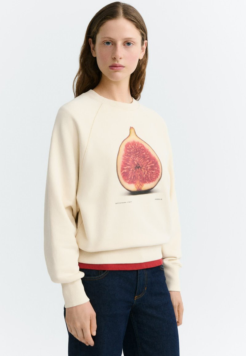 Jeune femme aux longs cheveux bruns porte un sweat crème avec une grande illustration détaillée d'un fruit de figue sur le devant.