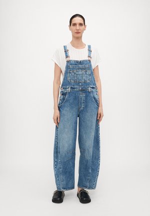 Salopette in denim blu chiaro, con bretelle regolabili, tasche frontali e gambe larghe con dettagli invecchiati sulle ginocchia. Indossata sopra una maglietta bianca.