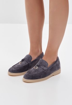 Voeten met grijze suède loafers met beige zolen en metalen lusdetail bovenop, staand op een witte ondergrond.