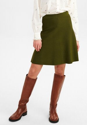 Femme portant une jupe trapèze vert olive, un chemisier blanc à fleurs, et des bottes en cuir marron montant jusqu'au genou, posant sur un fond blanc.