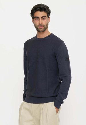 Uomo adulto con capelli scuri che indossa un maglione blu navy a maniche lunghe e pantaloni beige, in piedi davanti a uno sfondo bianco uniforme.
