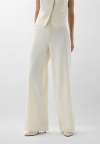 Costume crème clair avec un col ouvert, tissu texturé et pantalon à jambes larges. Comprend un design minimaliste et des chaussures élégantes à bout pointu.