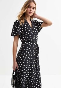 Vestido negro con lunares, de manga corta, con cuello, botones en la parte delantera y cintura atada. La tela parece suave y ligera.