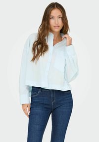 Light blue cropped button-up shirt with long sleeves, a single chest pocket, and buttoned cuffs, paired with dark blue denim jeans.