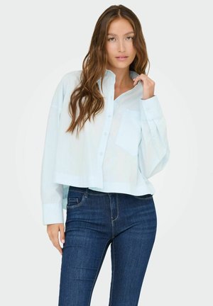 Camisa de botonadura en un azul claro, recortada y con mangas largas, un solo bolsillo en el pecho y puños con botones, combinada con jeans de mezclilla azul oscuro.
