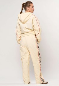 Nő, fonott hajjal, krémszínű melegítőben piros szegéllyel és logós csíkkal a nadrág oldalán, oldalra néz, kapucni nélkül.