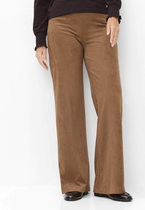 Personne portant un pantalon marron à jambes larges, des chaussures noires et un haut noir à manches longues, debout sur un fond blanc.