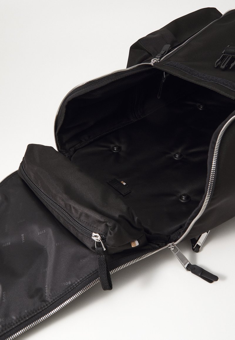 Sac à dos noir avec des ouvertures zippées, présentant un extérieur lisse, un intérieur doublé et une poche plate supplémentaire à l'intérieur.