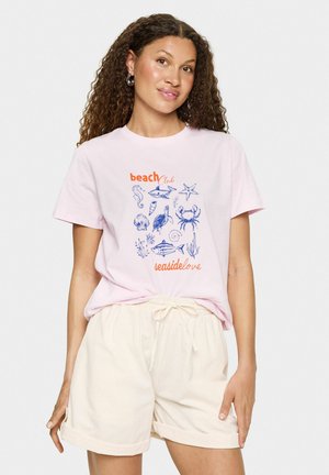 Camiseta de algodón rosa claro con gráficos marinos en azul, que incluyen un cangrejo y una estrella de mar, con el texto "beachClub" y "seasidelove". Combinada con pantalones cortos de color crema.