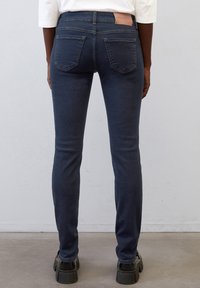 Slim-fit tmavě modré džíny vyrobené z denimu, s vysokým pasem, zadními kapsami a značkovou nášivkou na pase. Jsou obuty černé mohutné boty.