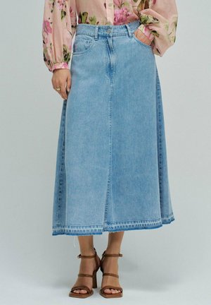 Mujer con falda midi vaquera azul clara, blusa floral de manga larga y sandalias marrones de tacón con tiras, de pie con una mano en el bolsillo.